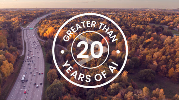 Greater Than 20 years of AI logo above a image taken from above where cars are driving on a highway next to a forest with fall colour leafs.