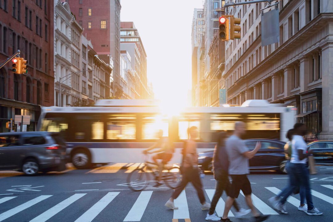 Busy urban intersection at sunset with a crowd of pedestrians crossing the street in the foreground, a cyclist riding through the crosswalk, and a city bus and cars in motion. Tall buildings frame the scene, and red traffic lights are visible on both sides.