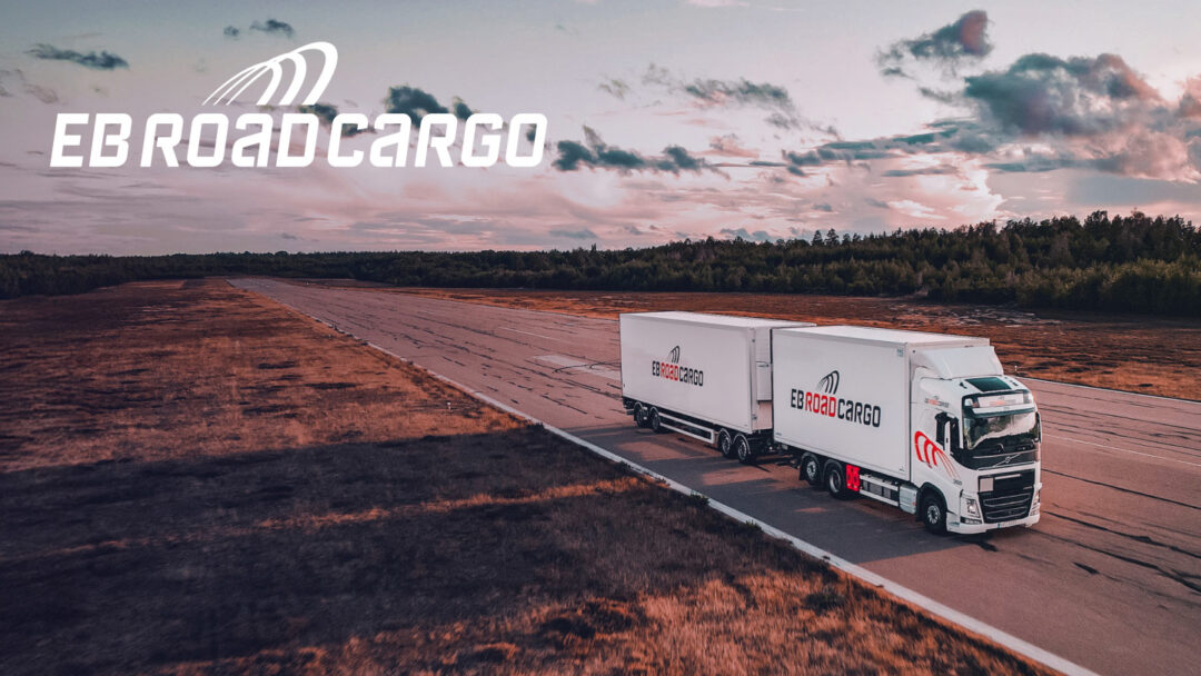 A truck branded with EB Road Cargo drives on a road in the sunset in an open landscape with forest around it and the EB Road Cargo logo to the left