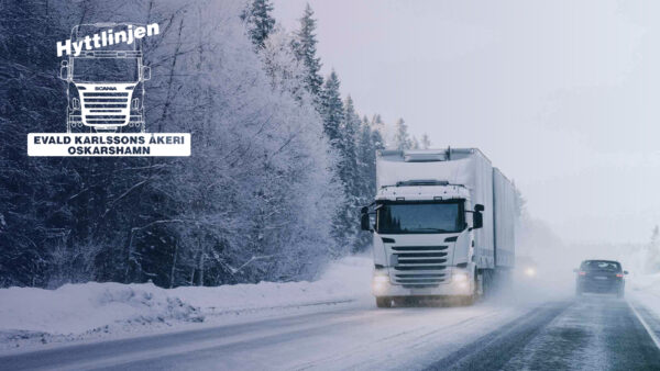 A white articulated truck driving on a winter road with snow and forest around. A passenger car is driving past it in the opposite direction. The Evald Karlsson Åkeri logo is in the upper left corner