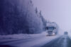 A white articulated truck driving on a winter road with snow and forest around. A passenger car is driving past it in the opposite direction.