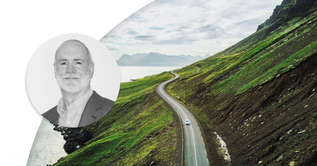 Car driving on the road next to a steep hill right by the water, mountains in the horizon. Profile image of Jim Noble.