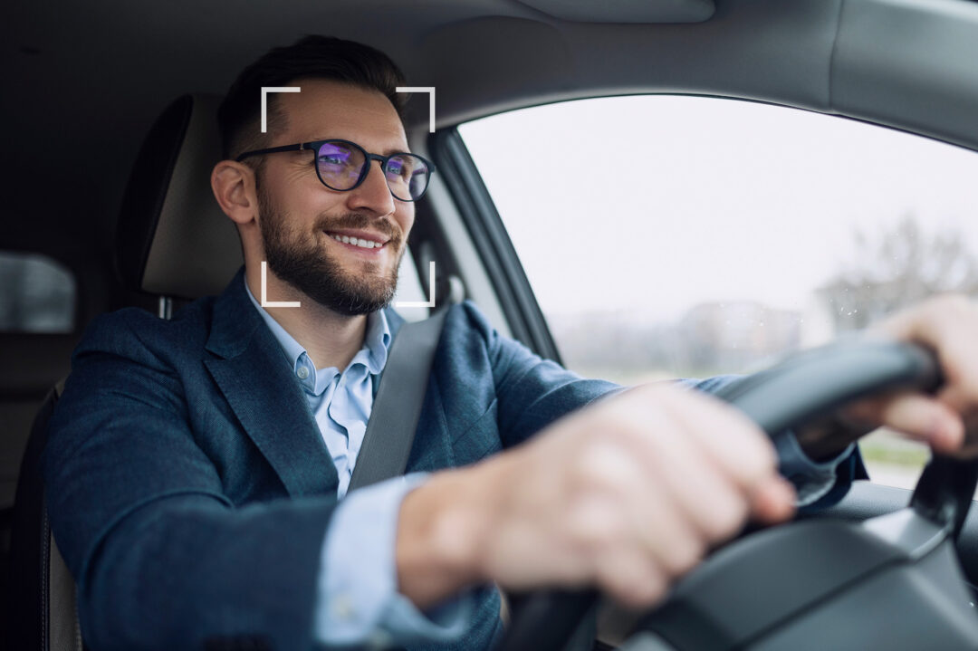 Close-up of a smiling man in business attire driving a car, wearing glasses and a seatbelt. A white square overlay highlights his face, suggesting facial recognition or driver monitoring technology.