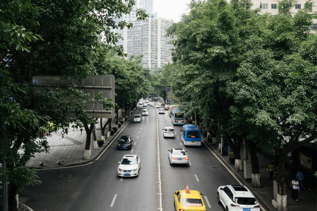 Tree-lined urban street with moderate traffic including cars, taxis, and blue buses traveling in both directions. Pedestrians walk along wide sidewalks beneath the trees, and high-rise apartment buildings are visible in the background.