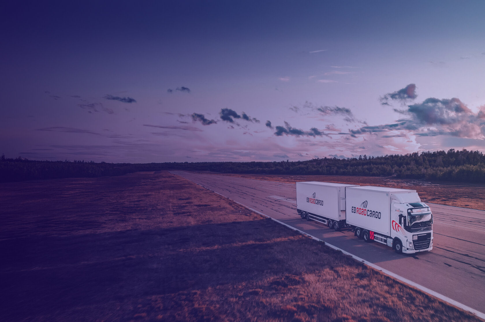 A truck branded with EB Road Cargo drives on a road in the sunset in an open landscape with forest around it