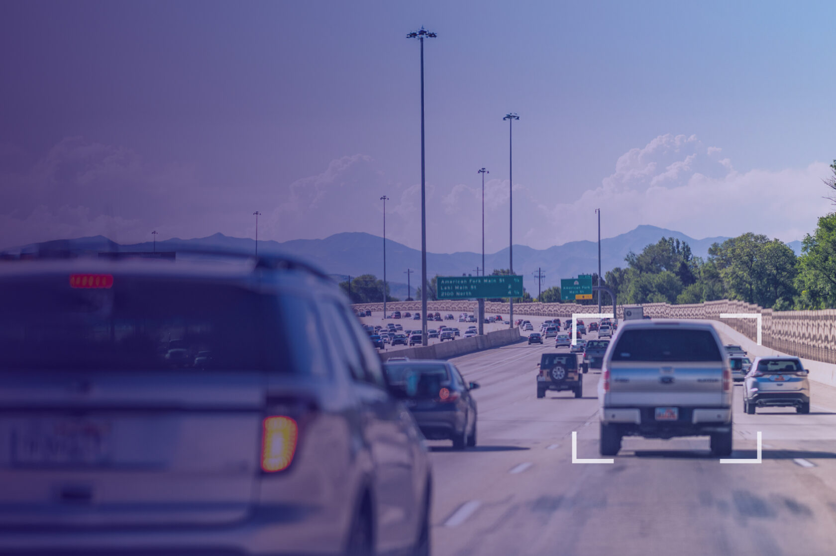 View of a busy multilane highway with several cars driving under clear skies, mountains visible in the distance. A white square overlay highlights a silver pickup truck in the center lane, suggesting vehicle tracking or monitoring technology.