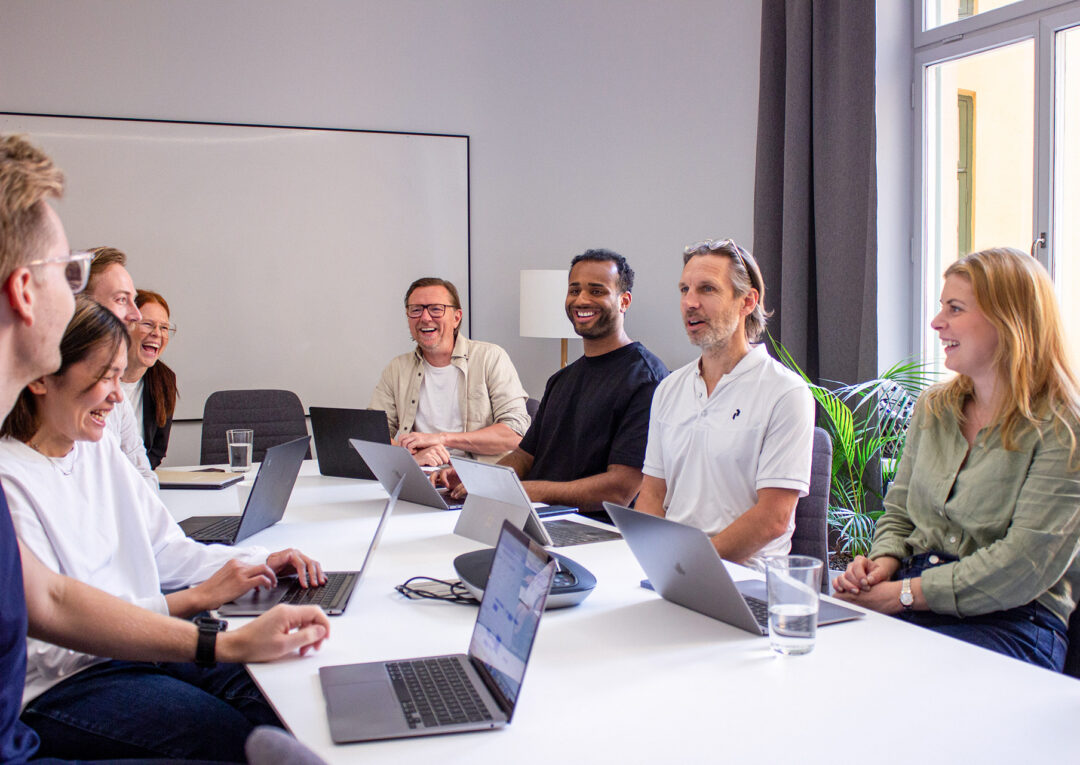 A group of eight colleagues smiling and laughing around a conference table during a meeting, each with a laptop in front of them, in a bright office with a large whiteboard and window in the background.