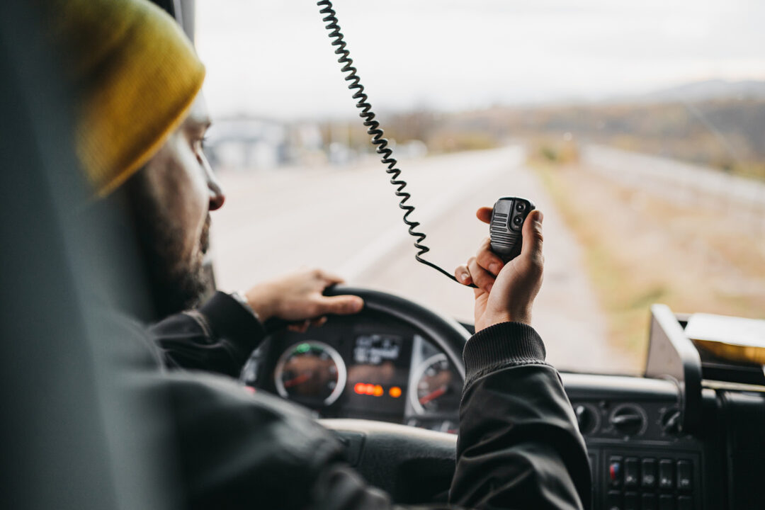 A male truck driver from behind with one hand on the steering wheel and one hand using the cb radio