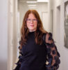 Liselott Johansson standing confidently in a modern office hallway, wearing glasses and a black top with sheer, decorative sleeves. She has long auburn hair and a composed expression.