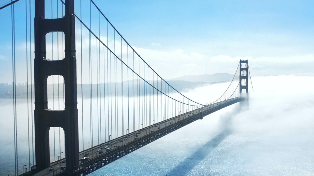 Suspension bridge above the water with low clouds around it and land in the horisont.