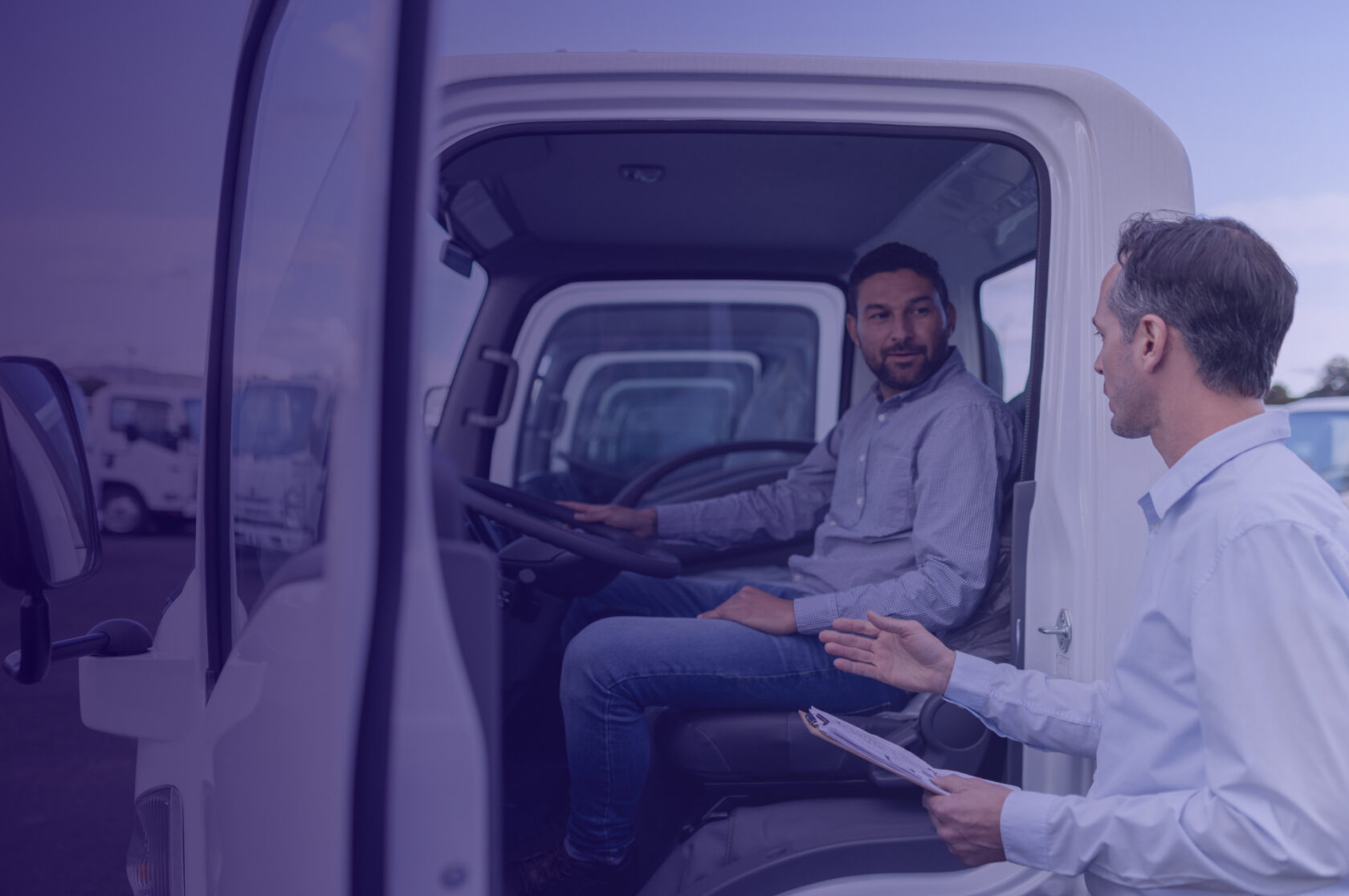 A man sitting in the driver’s seat of a truck receiving coaching from another man standing outside, both engaged in discussion, illustrating driver training and performance feedback.