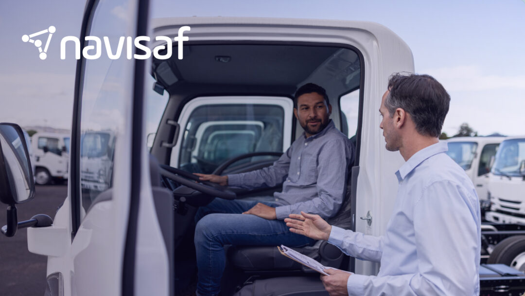 A man sitting in the driver’s seat of a truck receiving coaching from another man standing outside, both engaged in discussion, illustrating driver training and performance feedback. The Navisaf logo is in the upper left corner.