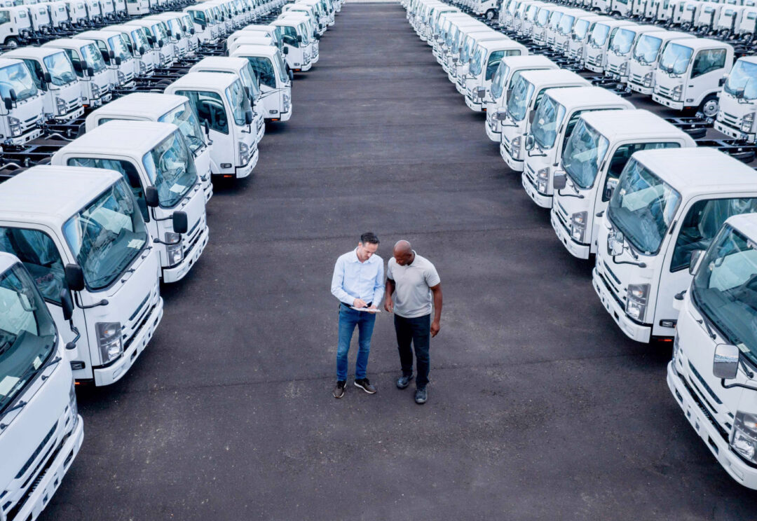 Two men standing between neatly aligned rows of white trucks in a fleet yard, reviewing a clipboard, symbolizing fleet management and inspection routines.