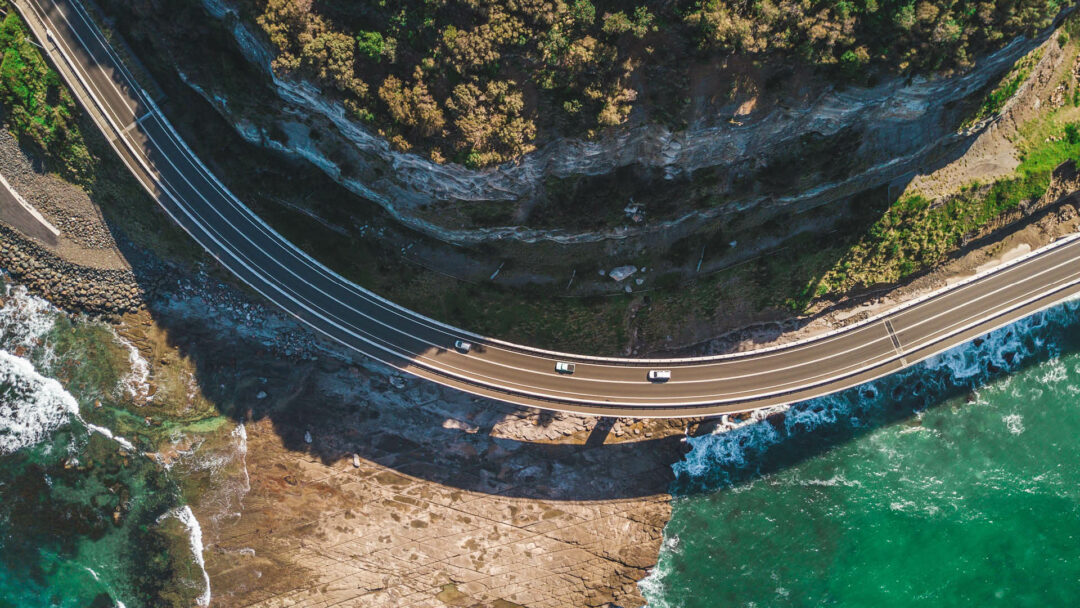 Road that winds along a mountain and the water's edge with three white cars driving in the same direction.