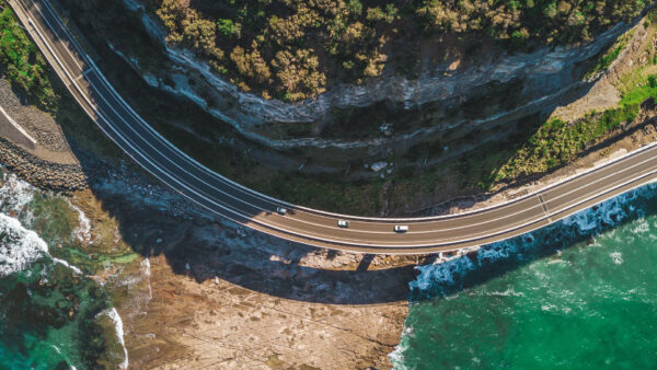 Road that winds along a mountain and the water's edge with three white cars driving in the same direction.