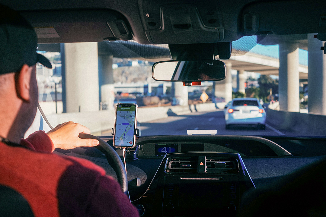 View from the back seat of a car showing a driver navigating through city traffic with a navigation app displayed on a smartphone mounted to the dashboard. Elevated highways and another vehicle are visible ahead through the windshield.