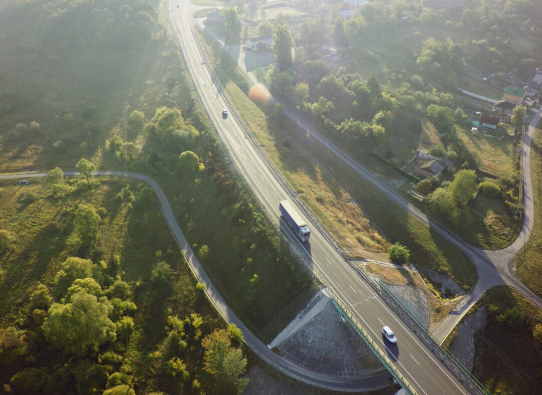Aerial view of a highway winding through a lush, green countryside at sunrise. A truck and several cars drive along the road, surrounded by forested hills, rural houses, and curving side roads, capturing the calm and efficiency of rural transport logistics.