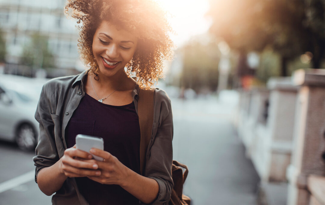 A woman walking on the street next to parked cars looking at her phone